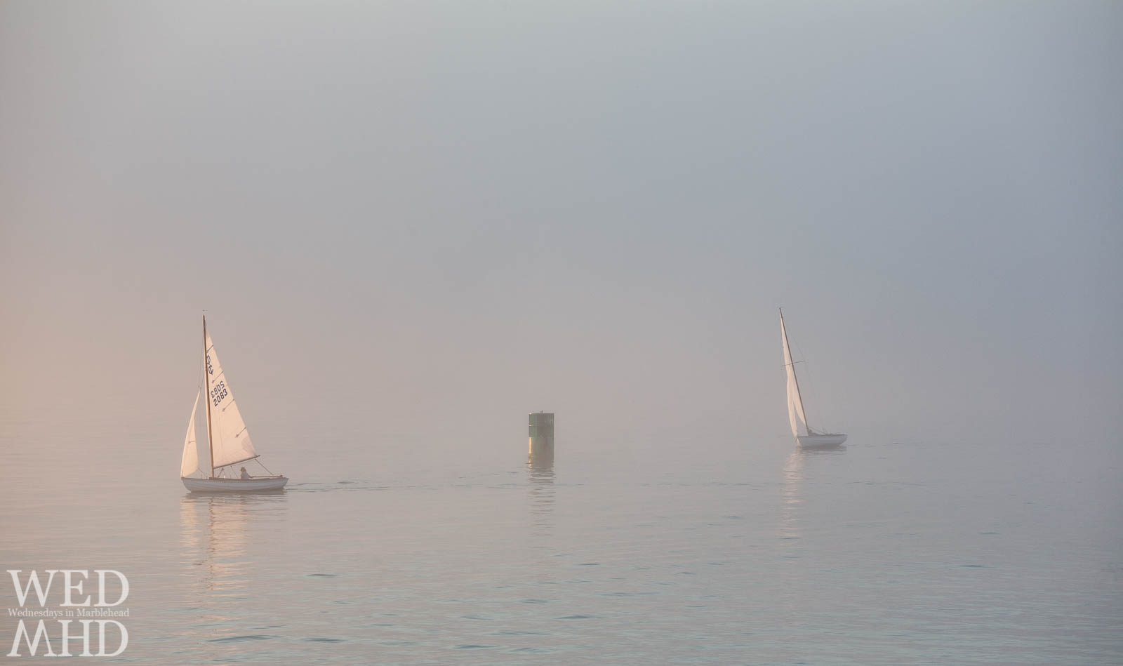 Two town class sailboats reflect sunlight as they are caught racing in the fog on a Summer evening