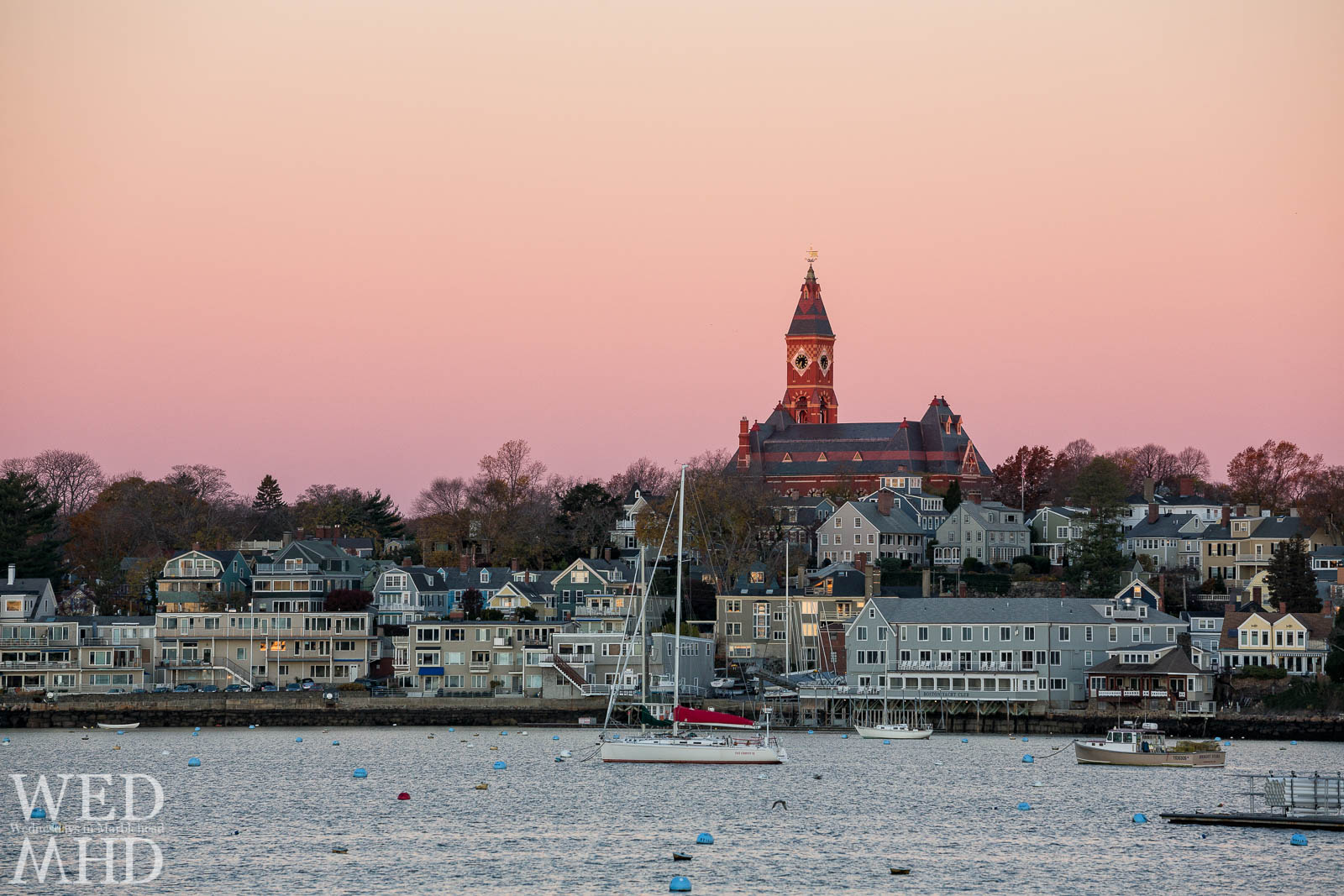 The boats Fast Company and Bright Stars remain in Marblehead Harbor on a mid-November morning with a pink-orange dawn over Abbot Hall