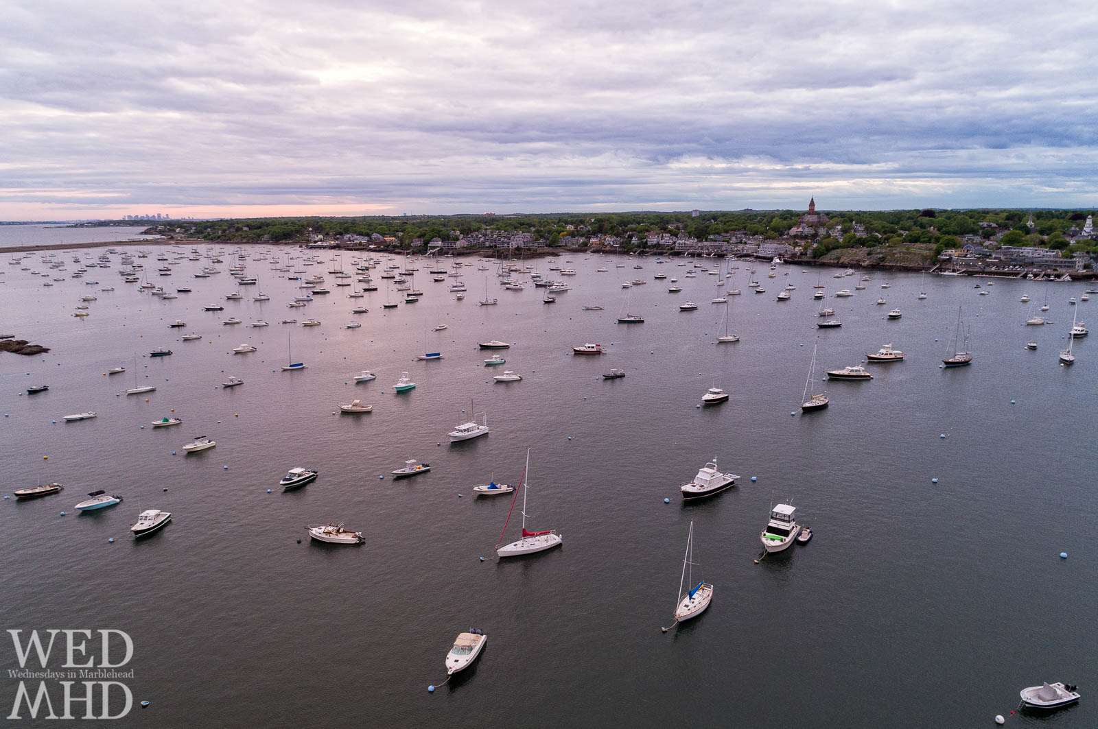 A faint sunset casts its glow on boats in the harbor as trees filled with foliage signal the arrival of Spring in Marblehead