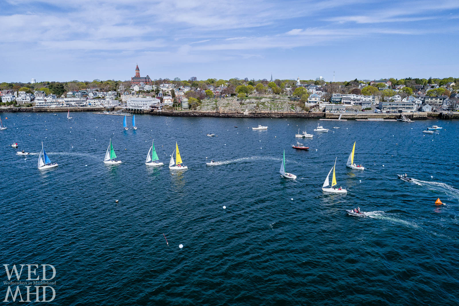 An aerial view of stiff competition during Jackson Cup racing in Marblehead Harbor features boats flying colorful sails as they cross in front of familiar landmarks