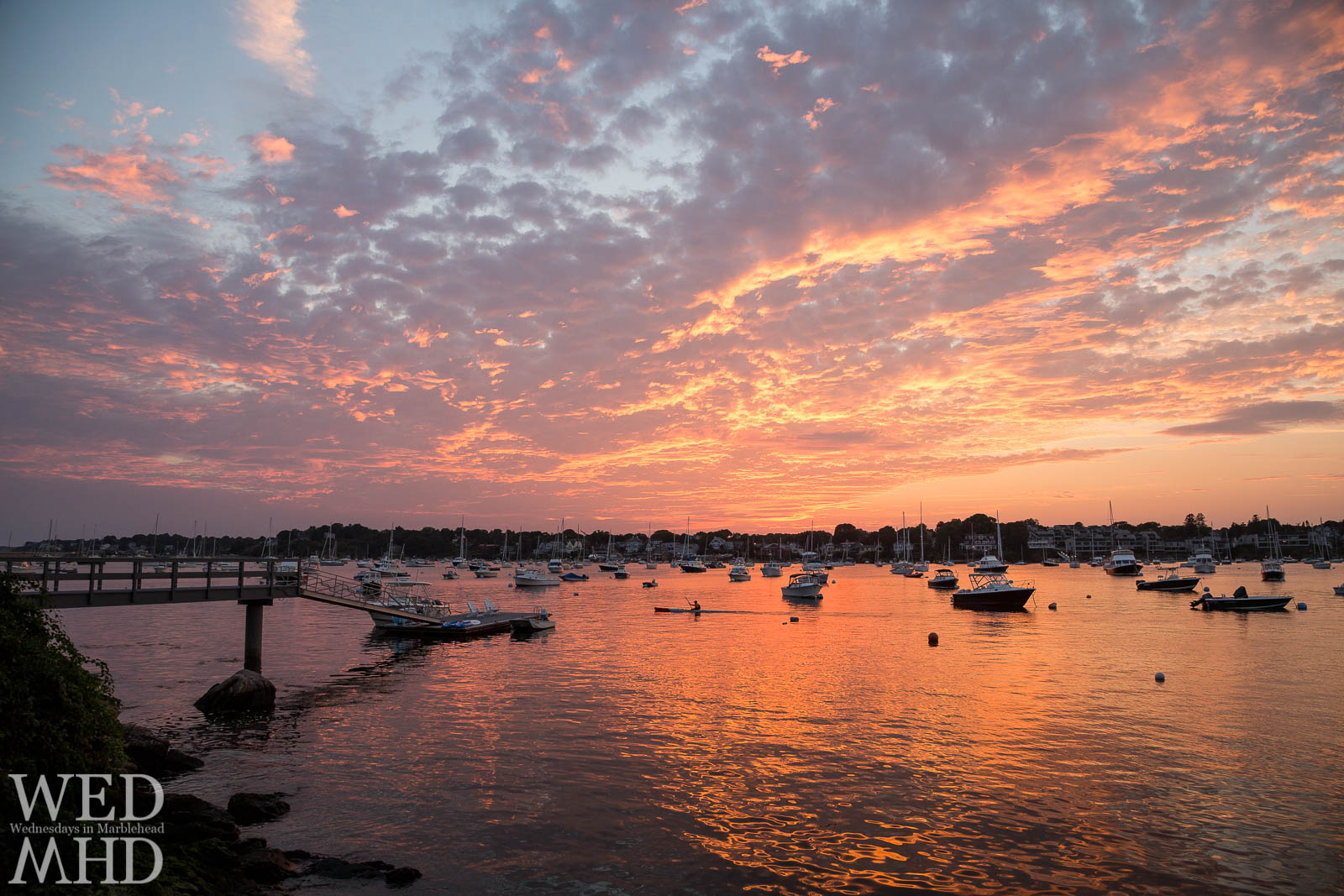 A man in a kayak is captured rowing through Marblehead Harbor as the sunset explodes above and reflects in the still water