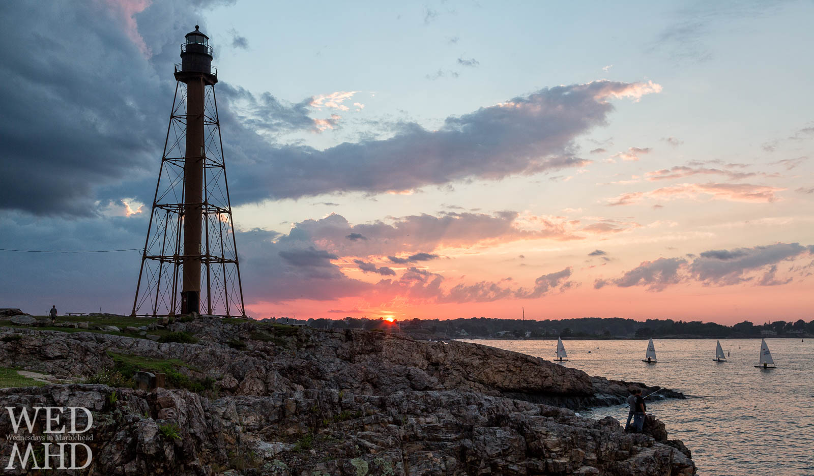 Four sailboats seen coming home as the sun dips below the horizon and paints the sky behind Marblehead Light