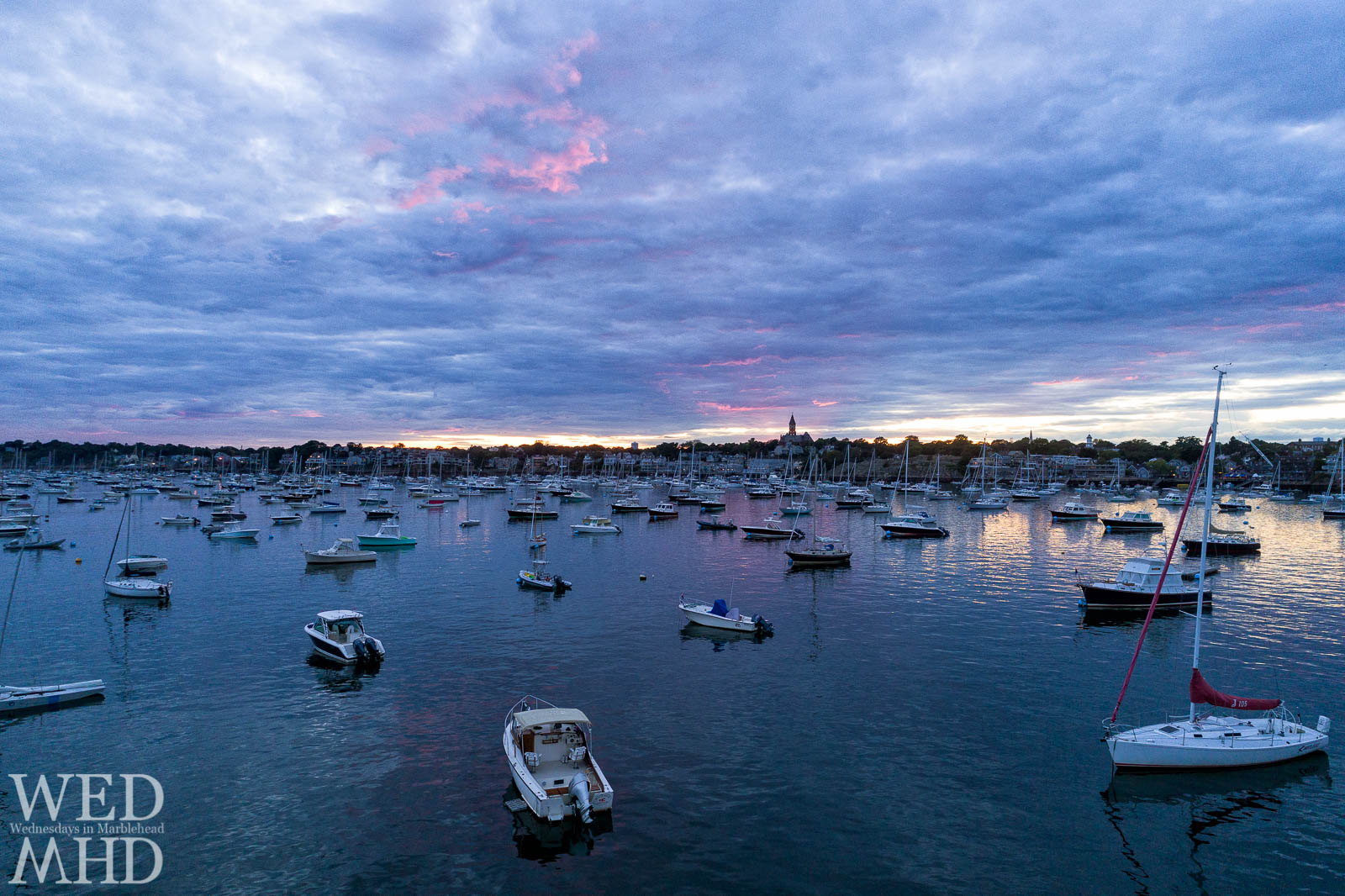 A low aerial view of a mid-harbor sunset reveals pink light behind Abbot Hall and high in the September sky as boats fill their moorings