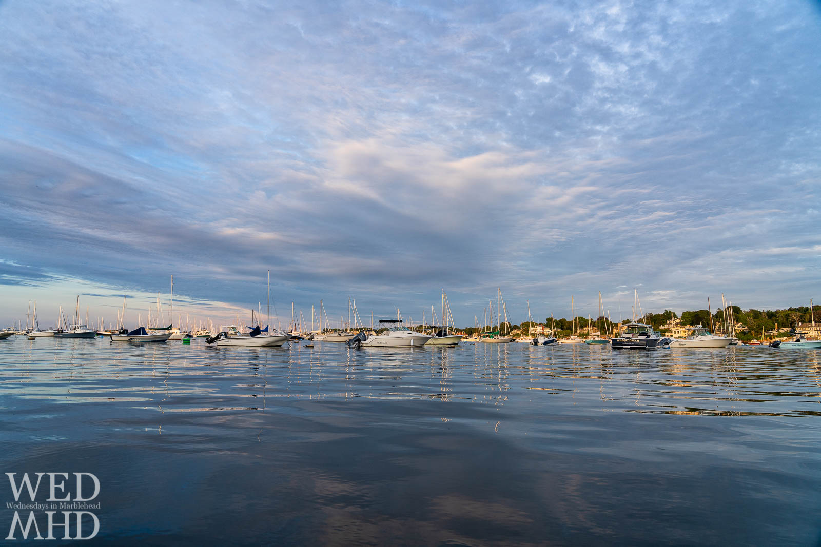 The still water of Marblehead Harbor creates perfect reflections of boats and their masts under a partly cloudy sky just before sunset on a September evening