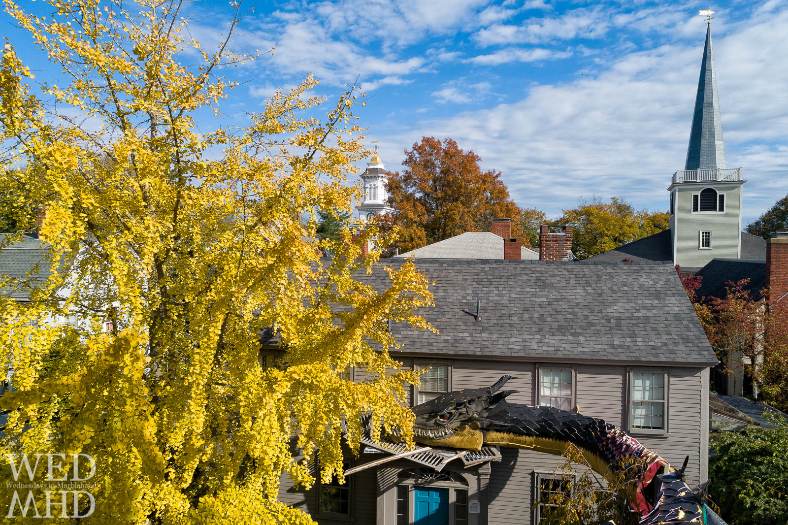 A dragon is seen resting on a house in Marblehead with two church steeples in the background