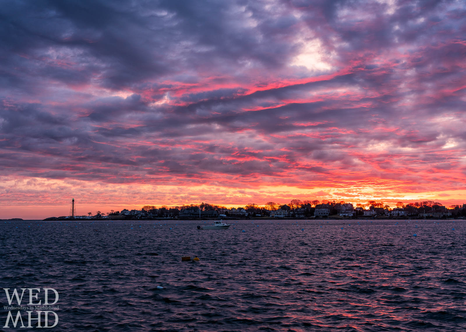 A brilliant sunrise paints the sky in pink and orange over a purple harbor with Marblehead Light's green beacon shining bright