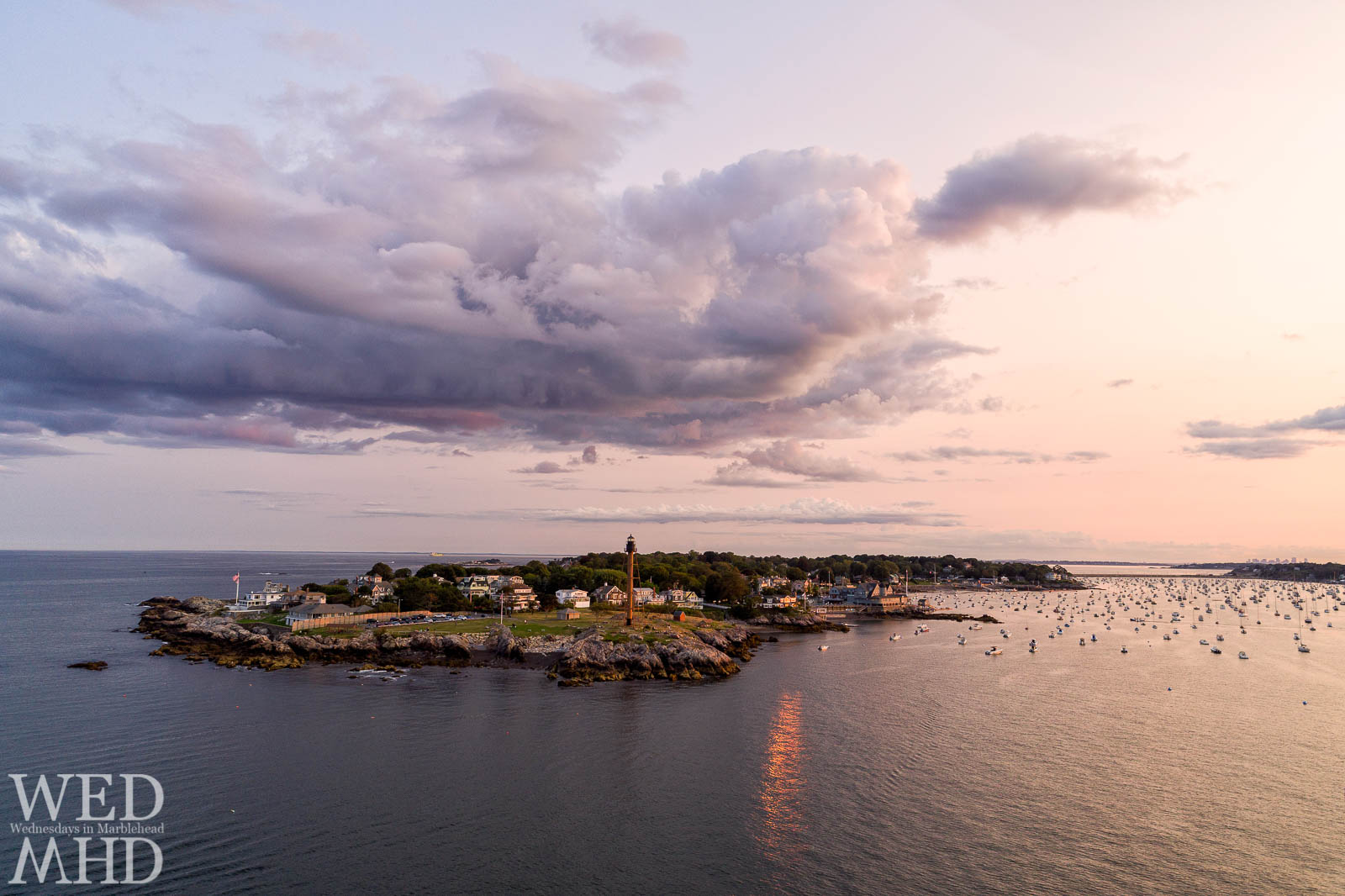 An aerial image captures the setting sun reflecting off a house on the Neck with Marblehead Light standing tall over the homes and foliage