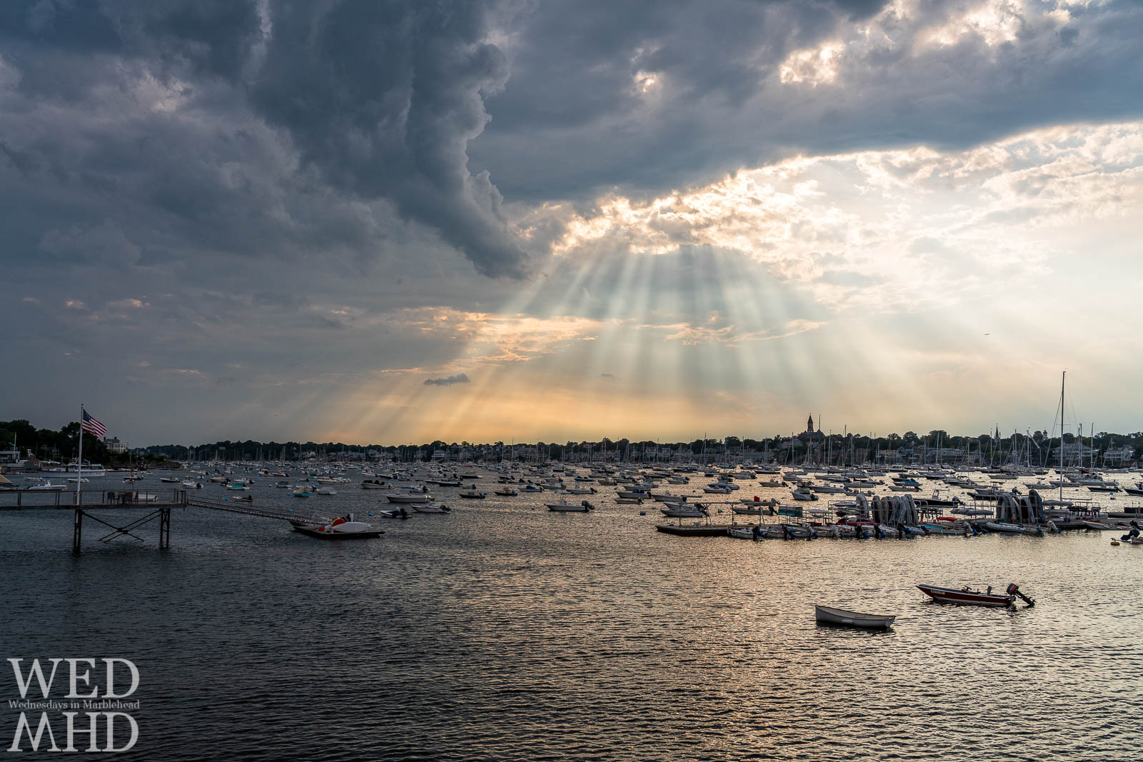 A full harbor and a flag flying at the end of a pier on the Neck are bathed by Marblehead Rays pouring through broken cloud cover