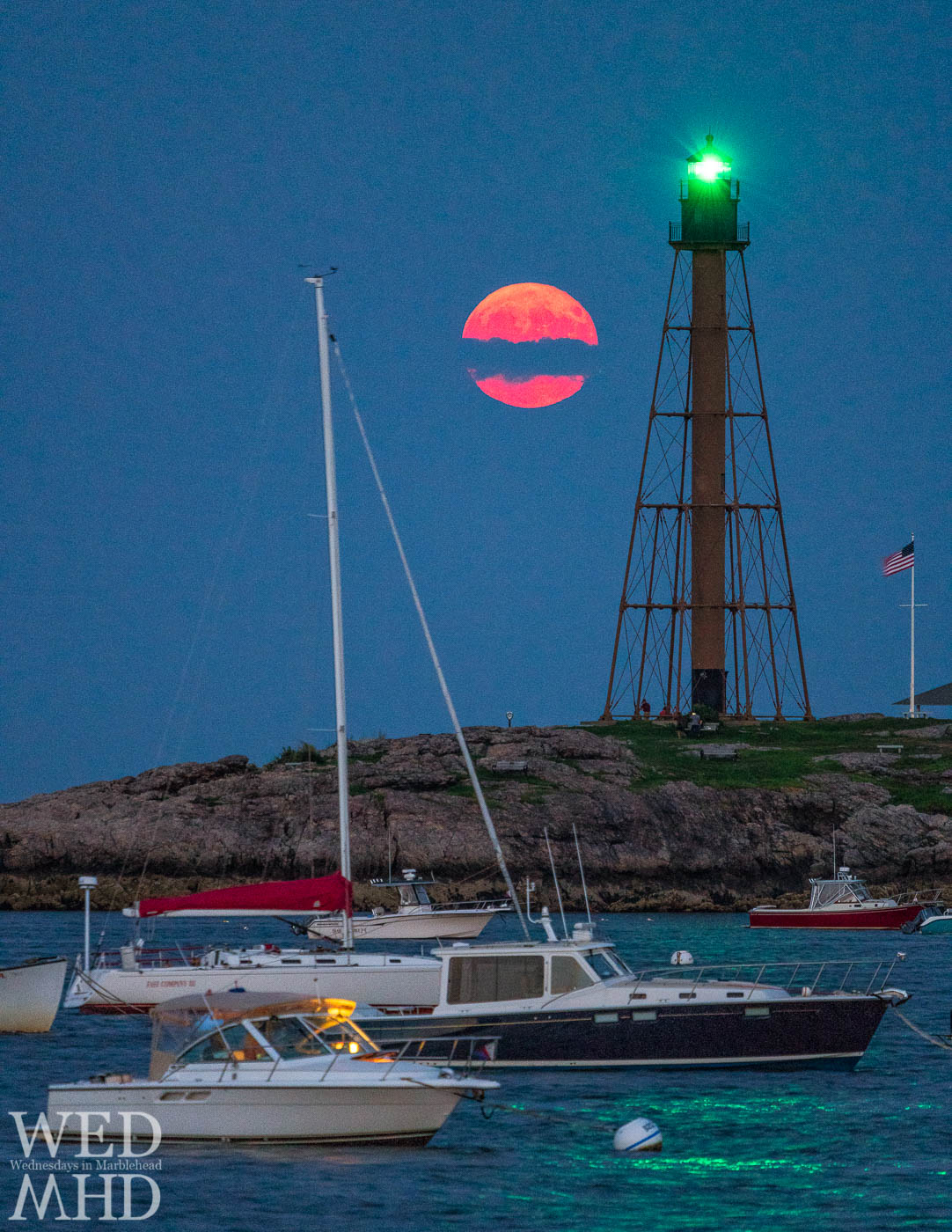 The moon rises through a band of clouds making it appear broken in half against the green glow of Marblehead Light