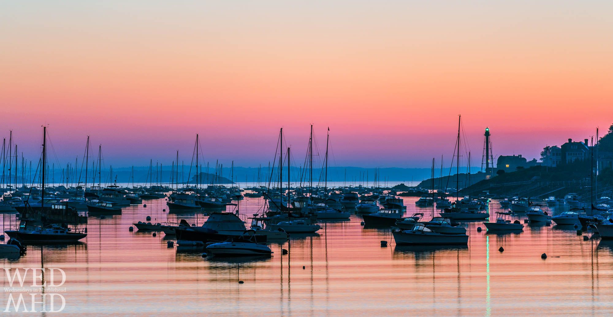 Dawn colors reflect off the water of Marblehead Harbor as does the lighthouse's green beacon while a single light signals an early riser on the Neck