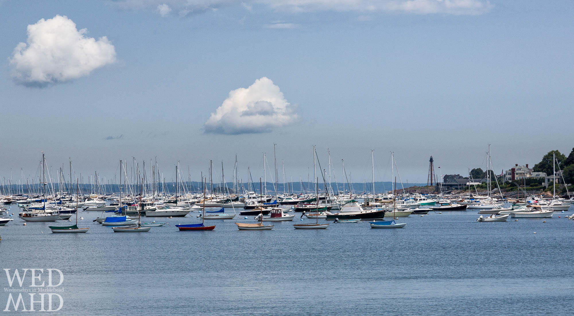 Two clouds over a full Marblehead harbor with town class sailboats in the foreground and Marblehead Light standing tall