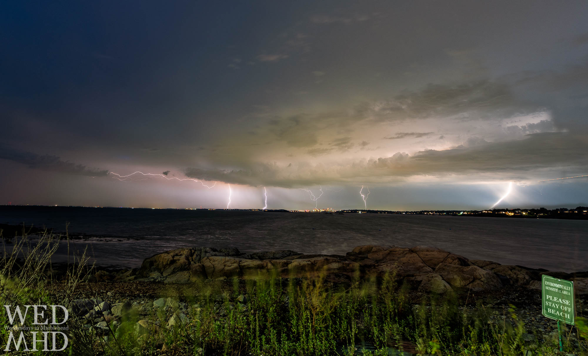 Lightning strikes over Boston as seen from a stretch along Ocean Ave on Marblehead Neck with a sign asking to please stay off beach